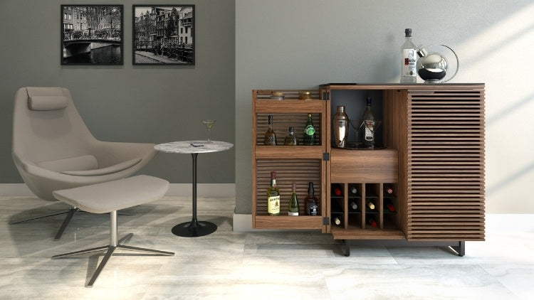 Modern wooden cabinet with horizontal slats and black base used as a liquor cabinet in a room with chair and table. 