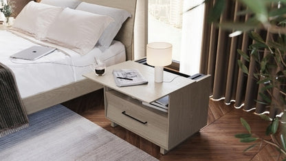 28" KAVA nightstand in washed oak in bedroom with top slid open, with notebook, drink, charging laptop, and lamp.