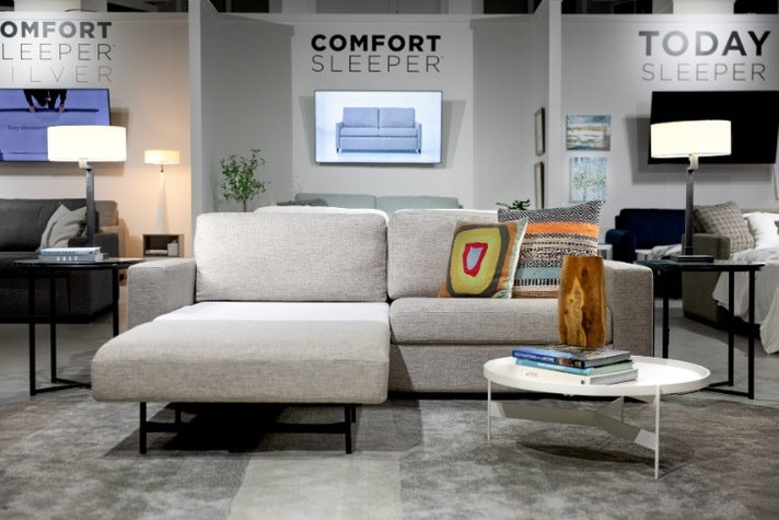 Gray sectional sofa with decorative pillows in a showroom setting, featuring 'Comfort Sleeper' branding.
