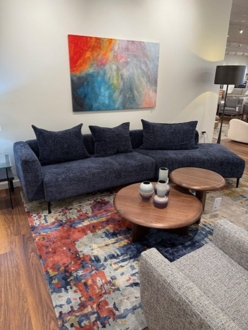 A navy blue angle sectional sofa with throw pillows, placed on a multicolored area rug in a showroom setting with a painting on the wall and a round wooden coffee table in the foreground.