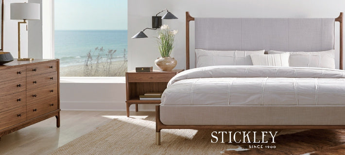 Stickley bedroom furniture set featuring a wooden bed frame with a light gray upholstered headboard, dressed in white bedding, shown in a bright coastal-style bedroom with a dresser, nightstand, and ocean view.