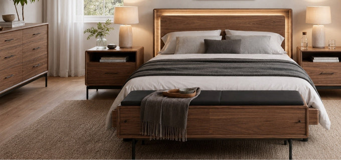 A modern bedroom scene featuring a wooden bed frame with an integrated warm LED backlight in the headboard, dressed in white and gray bedding, with a matching wooden bench at the foot of the bed.