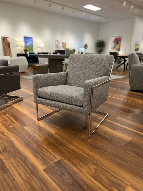 A gray upholstered Hi-Wire chair with a polished stainless steel base, positioned in a showroom setting.